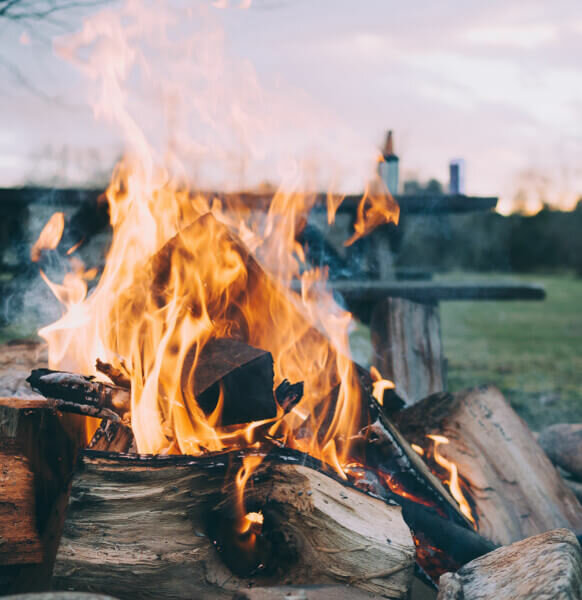 Biike Arrangement | Severin*s Resort and Spa Gemütliches Lagerfeuer im Freien, umgeben von Holzscheiten, vor einer weiten, grünen Landschaft bei Sonnenuntergang.