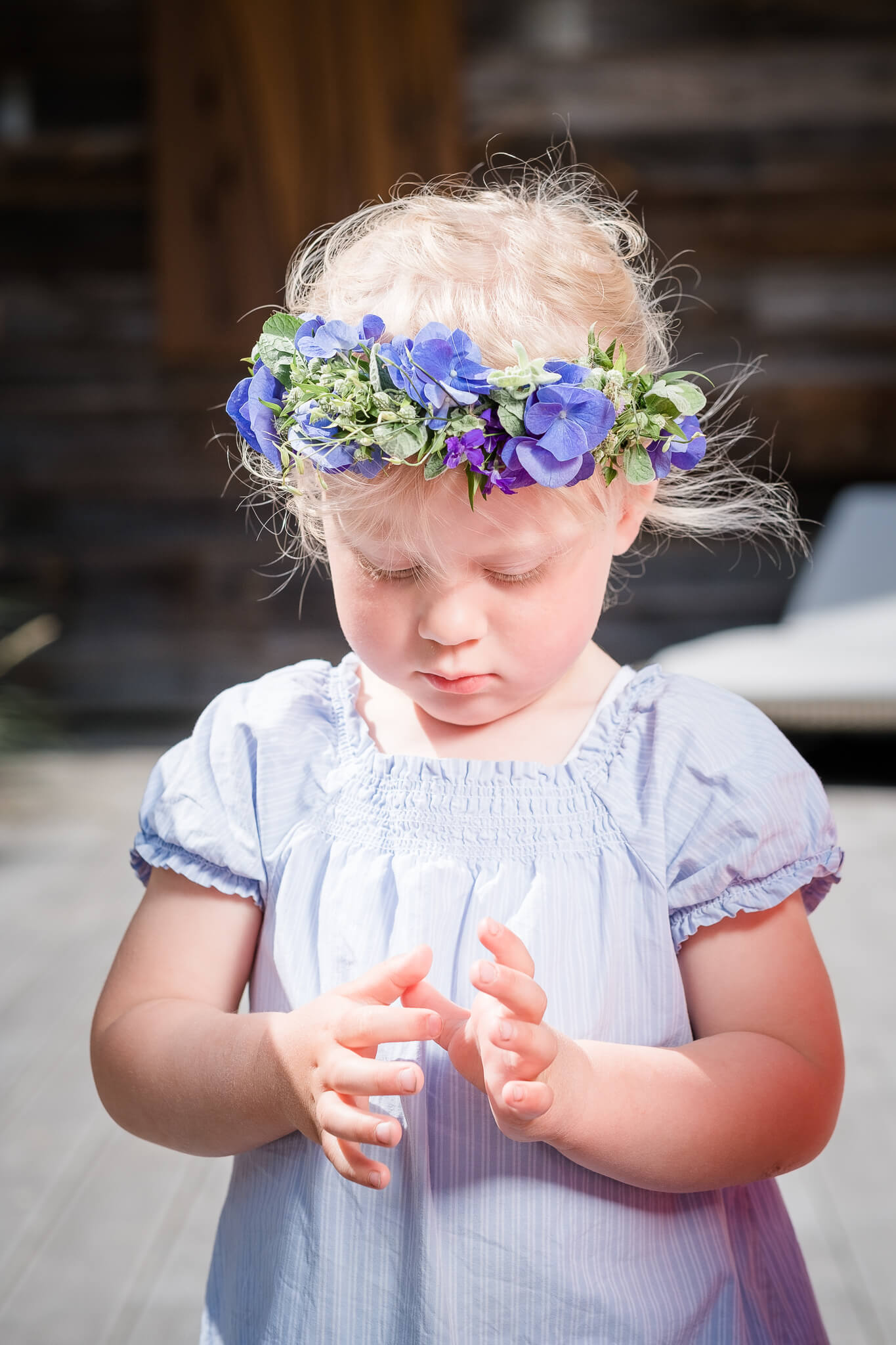 Kleines Kind mit Blumenkranz und blauem Kleid im Sonnenlicht vor Holzwand im Severin*s Resort & Spa auf Sylt.
