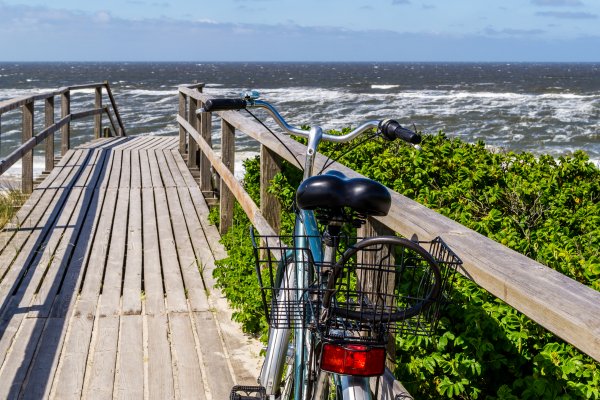 Fahrrad an Holzsteg mit Blick auf die raue Nordsee, umgeben von grünen Büschen, bei sonnigem Wetter.