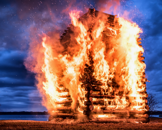 Ein brennendes Holzhaus am Abend, umgeben von dramatischem Himmel und Funkenflug.
