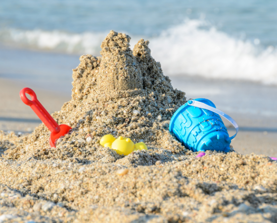 Sandburg am Strand mit roter Schaufel und blauem Eimer, Meer im Hintergrund.