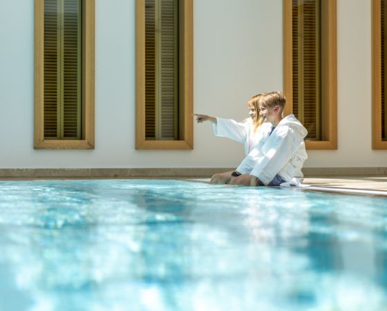 Zwei Kinder sitzen in Bademänteln am Pool-Beckenrand, ein Kind zeigt in die Ferne - Severin*s Resort & Spa