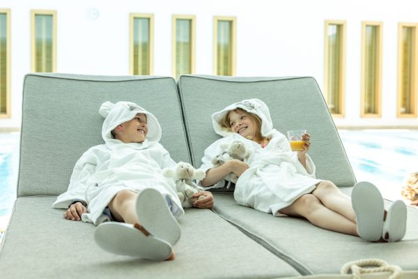 Zwei Kinder liegen in weißen Bademänteln auf einer Liege. Ein Kind hält ein Getränk in der Hand, sie lachen sich an - Severin*s Resort & Spa