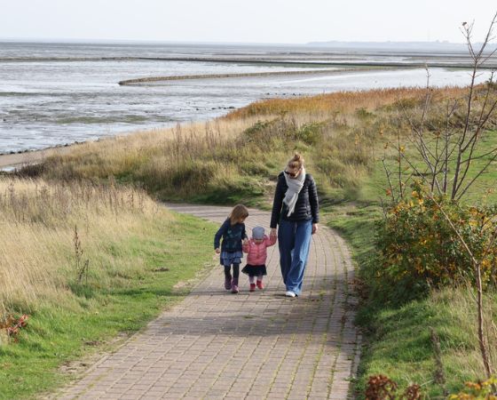 Mutter und Kinder laufen Hand in Hand einen Weg entlang, im Hintergrund sieht man Dünen und das Meer - Severin*s Resort und Spa