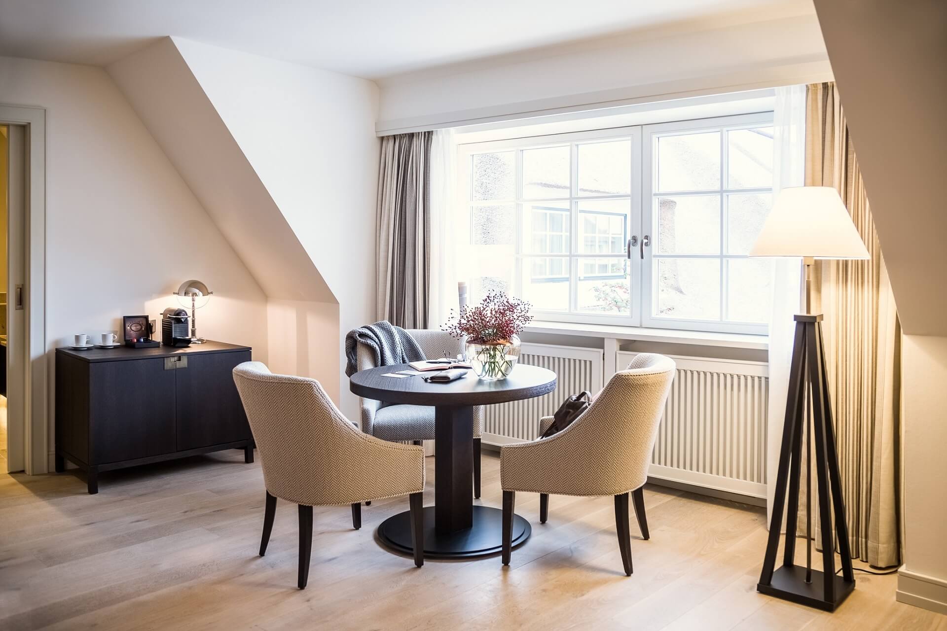 Gemütliche Sitzecke mit rundem Tisch, zwei Sesseln, großer Fensterfront und stilvoller Stehlampe im Severin*s Resort & Spa.