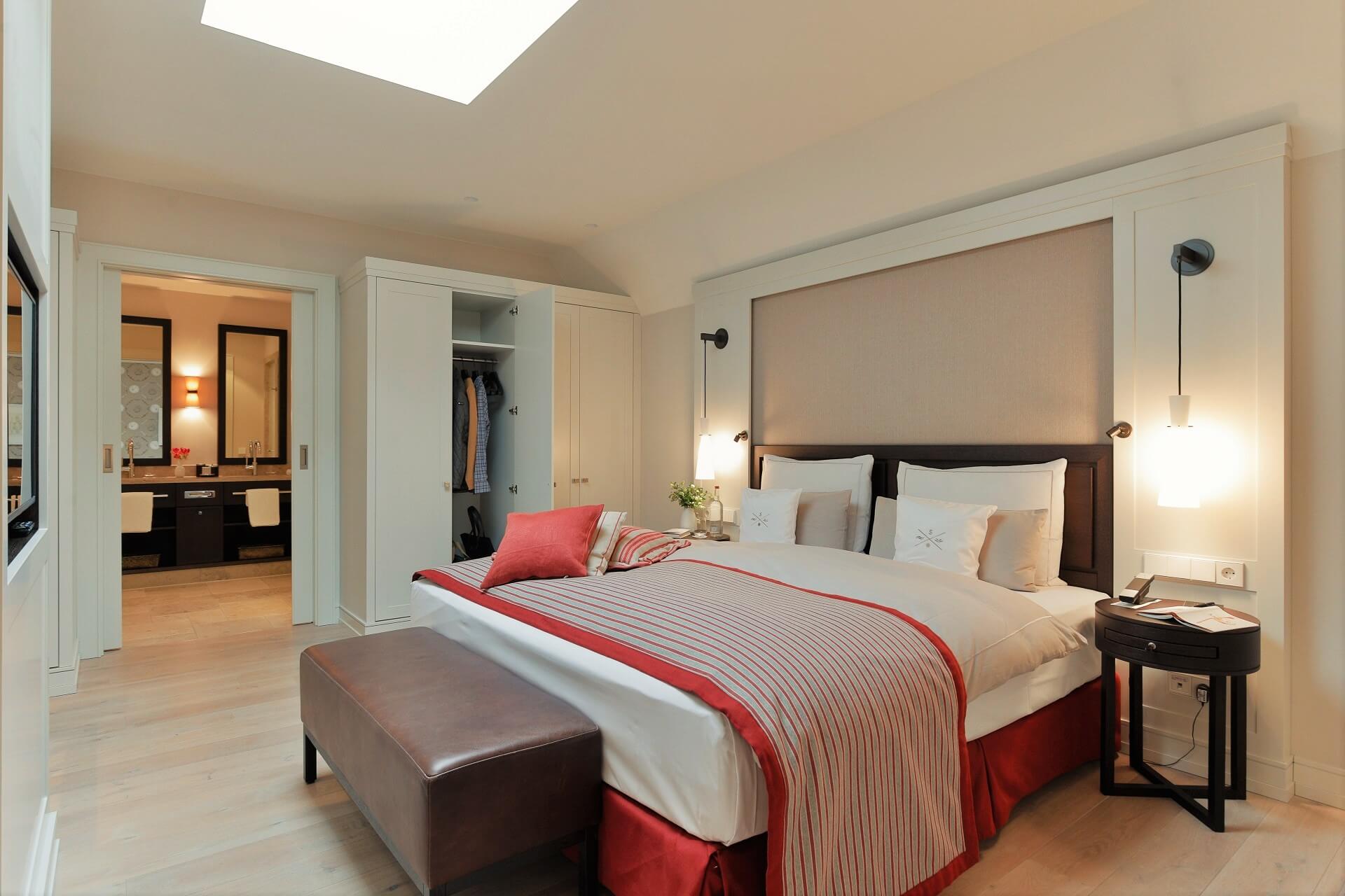 Elegantes Hotelzimmer mit Doppelbett, roten Akzenten, Sitzbank, offener Garderobe und Blick ins stilvolle Badezimmer.