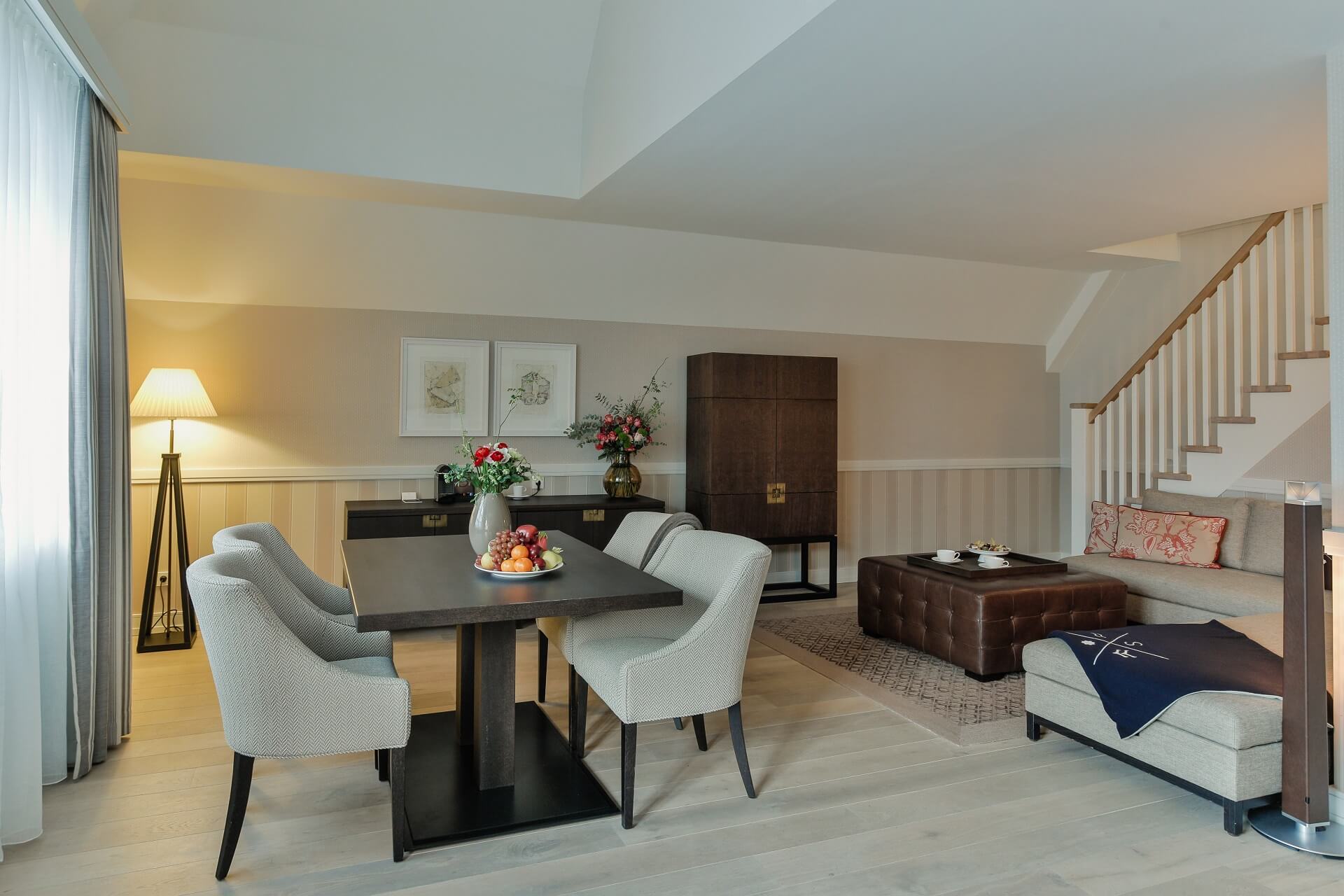 Elegantes Wohnzimmer im Severin*s Resort mit Esstisch, bequemen Sesseln, Sofa, Blumen und Treppe.