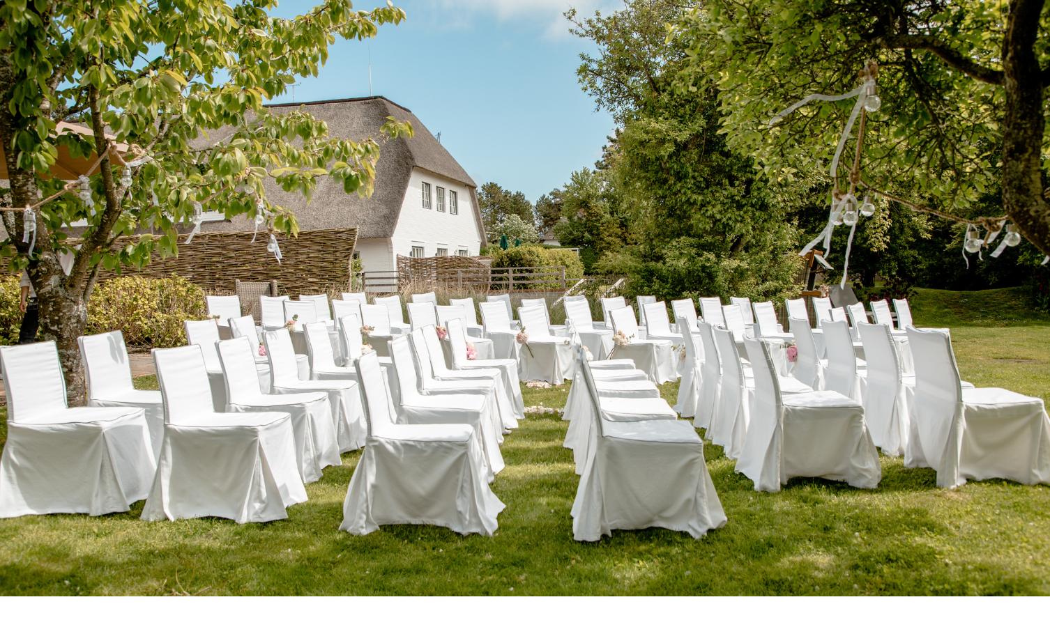 Weiße Stühle im Freien, für eine Hochzeit arrangiert, umgeben von Bäumen und einem traditionellen Reetdachhaus.