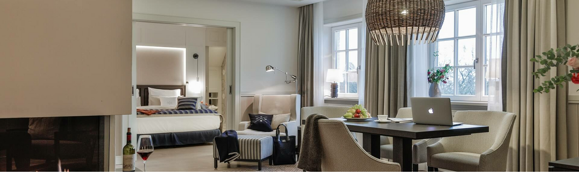Elegant hotel room with fireplace, cozy seating area, dining table, modern decor and large bed in the background.