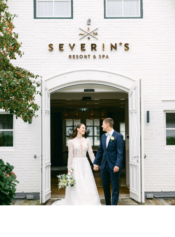 A smiling bride and groom stand in front of the entrance to Severin*s Resort & Spa on Sylt.