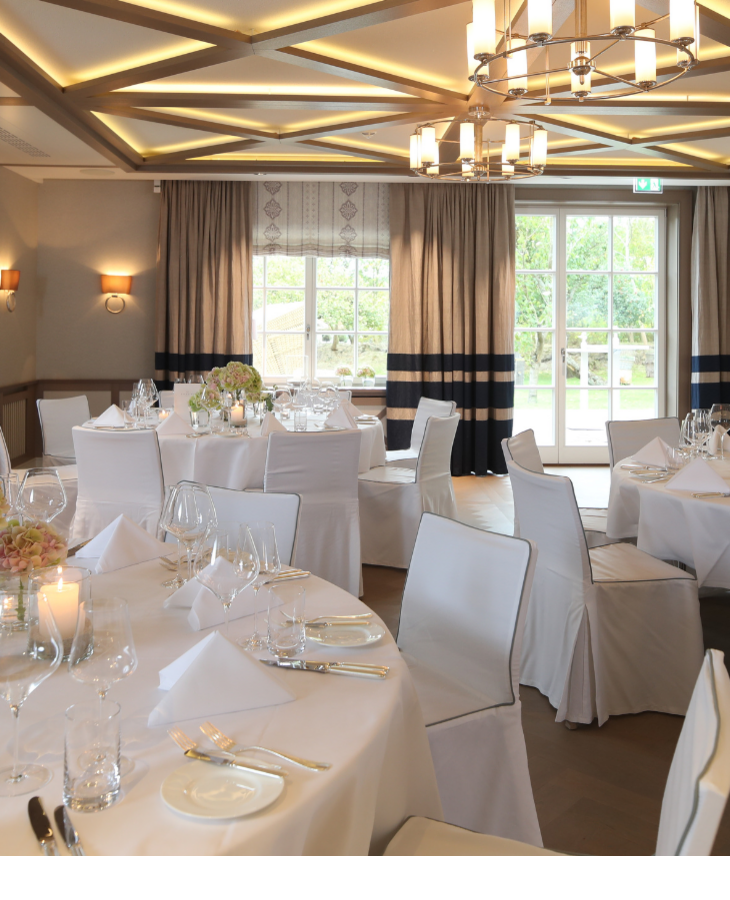 Eleganter Bankettsaal mit runden, weiß gedeckten Tischen, Kronleuchter und Blick auf den Garten durch große Fenster.