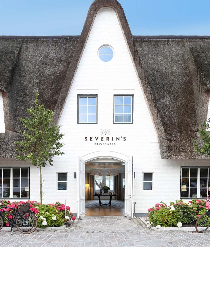 Reetgedecktes Hotel Severin*s auf Sylt mit blühenden Hortensien und Fahrrädern vor dem Eingang.