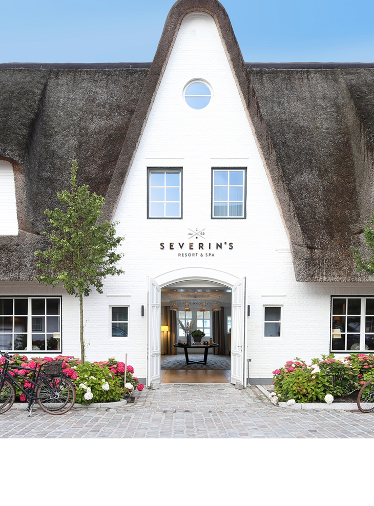 Reetgedecktes Hotel Severin*s auf Sylt mit blühenden Hortensien und Fahrrädern vor dem Eingang.