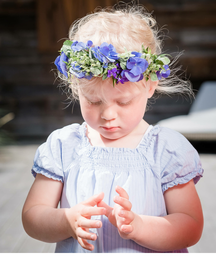 Kind mit Blumen in den Haaren Kleines Kind mit Blumenkranz und blauem Kleid im Sonnenlicht vor Holzwand im Severin*s Resort & Spa auf Sylt.