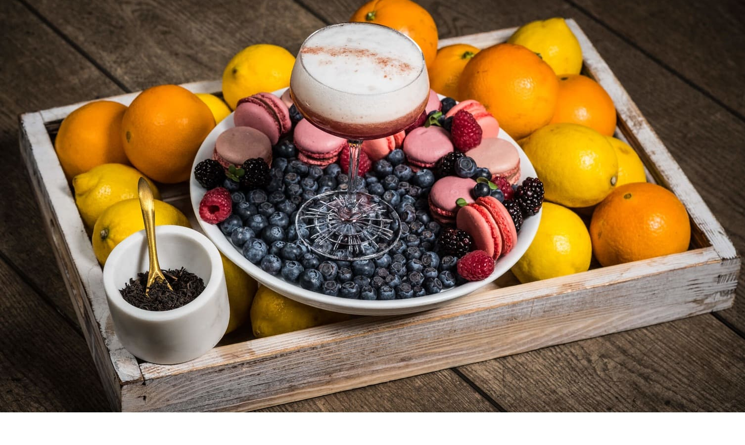 A bowl of macarons, berries and a cocktail surrounded by oranges and lemons on a wooden table.
