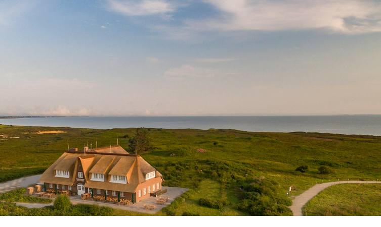 Landhaus Severin*s Morsum Kliff Reetgedecktes Hotel in grüner Landschaft mit Meerblick im Hintergrund bei Sonnenuntergang.