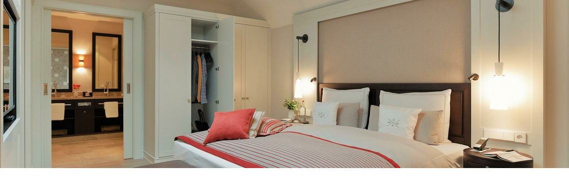 Elegant hotel room with double bed, red accents, bench, open wardrobe and view of the stylish bathroom.