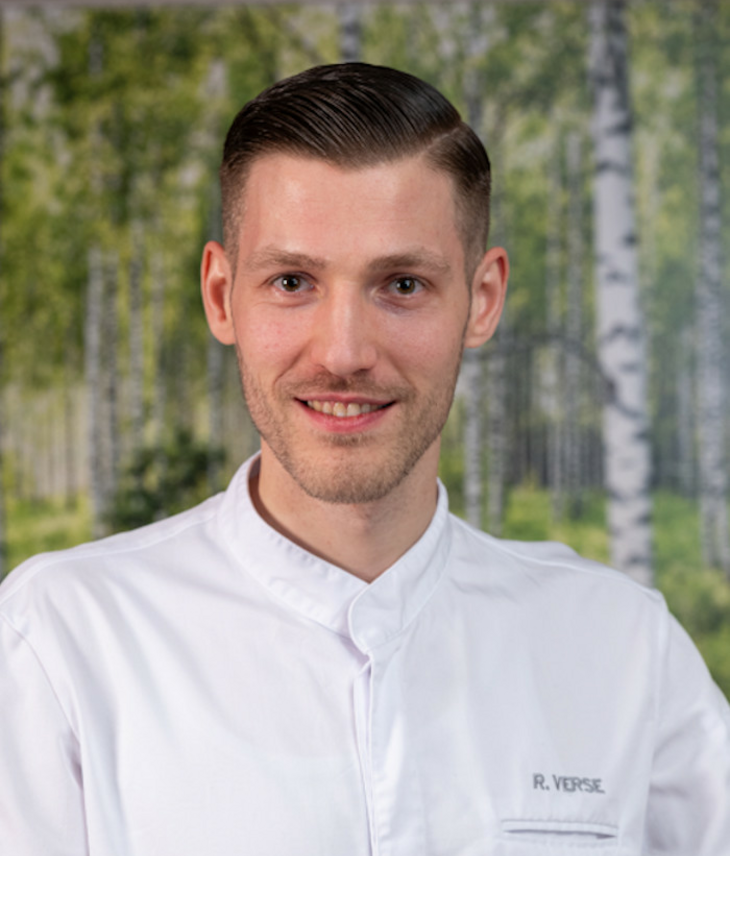 A chef in a white uniform smiles in front of a background with a birch forest motif.