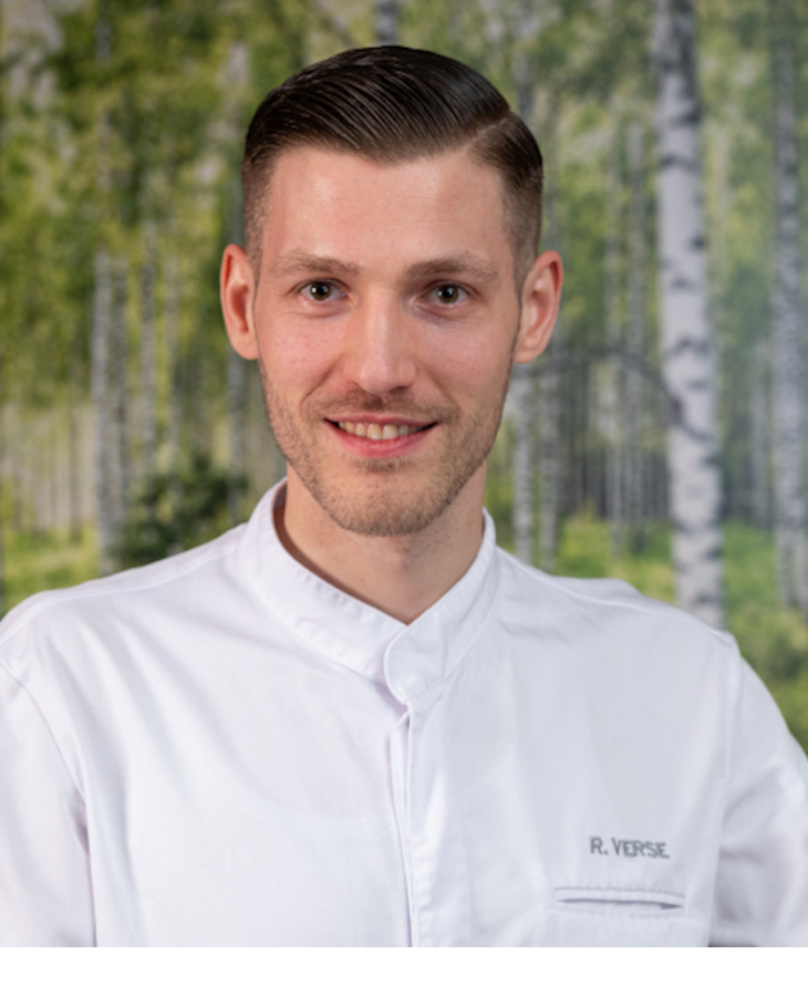 Ein Koch in weißer Uniform lächelt vor einem Hintergrund mit Birkenwaldmotiv.