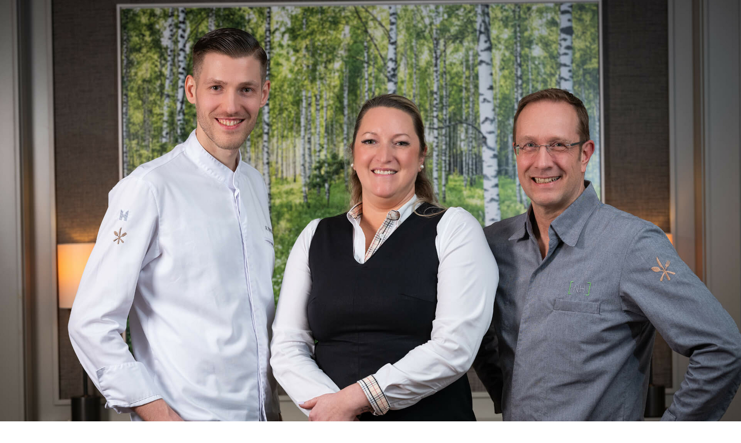 Drei lächelnde Hotelmitarbeiter in Uniform vor einem Bild mit Birkenwald im Severin*s Resort & Spa auf Sylt.