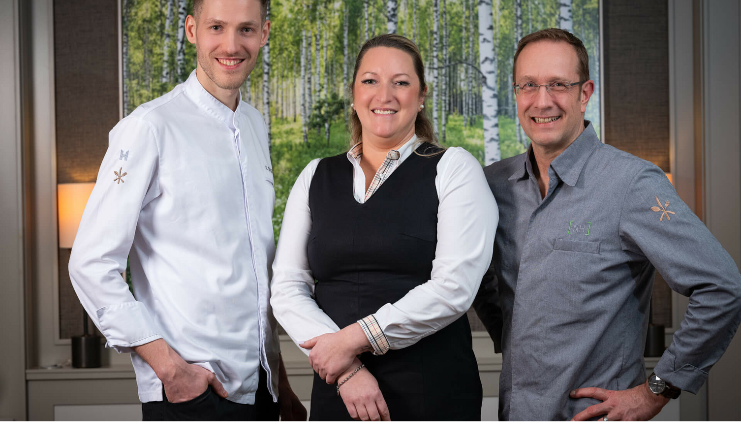 The Tipken`s by Nils Henkel team on Sylt Three smiling hotel employees in uniform in front of a picture of a birch forest at Severin*s Resort & Spa on Sylt.