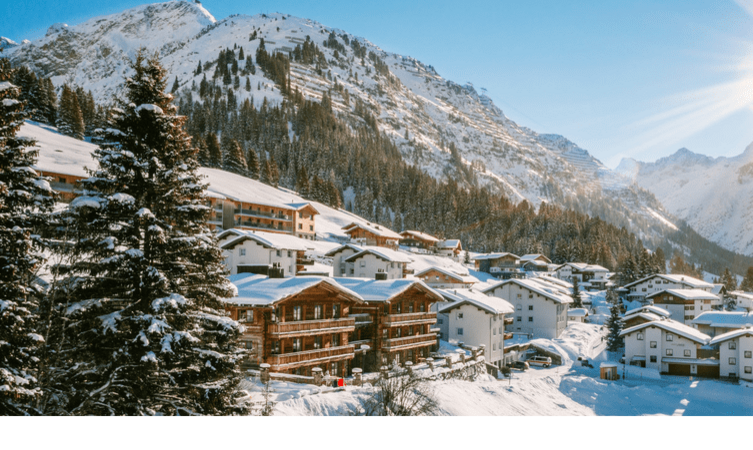 A snow-covered mountain landscape with chalet houses under a blue sky, surrounded by conifers.