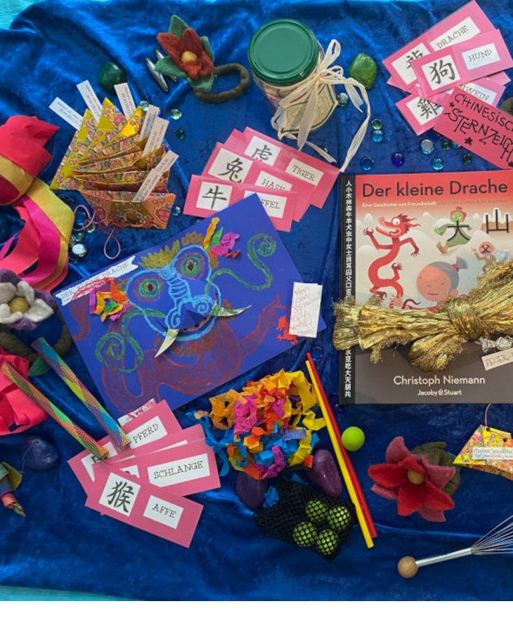 Buntes Arrangement mit Buch "Der kleine Drache", Bastelmaterialien und chinesischen Sternzeichen auf blauem Hintergrund.