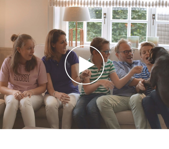 Several people are sitting together on a sofa in a bright living room; a play symbol indicates a video.