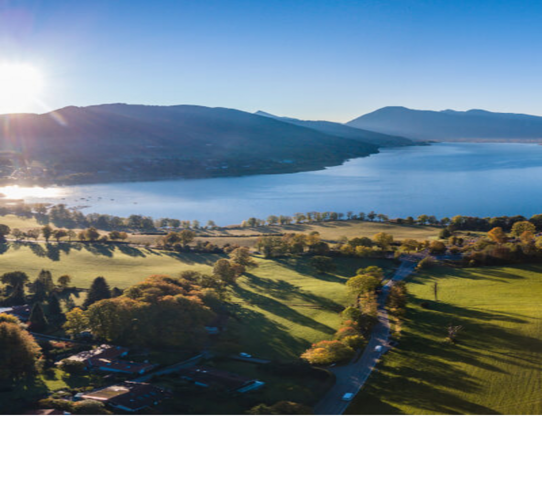 Luftaufnahme eines Sees mit umliegenden grünen Wiesen und Bergen im Hintergrund bei Sonnenaufgang.