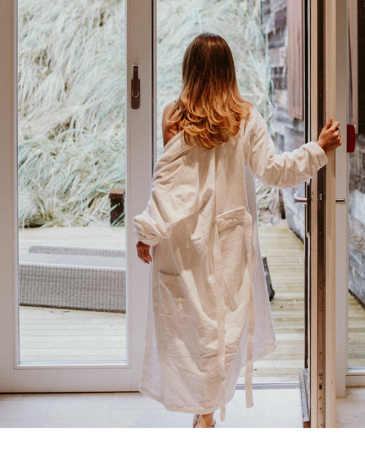 Woman in a bathrobe opens the patio door, looks out over the wooden deck and dunes at Severin*s Resort & Spa Sylt.