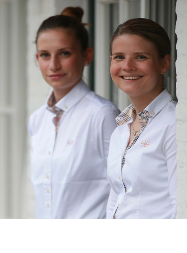 Zwei lächelnde Hotelmitarbeiterinnen in weißen Uniformen stehen vor einer hellen Wand des Severin*s Resort & Spa.