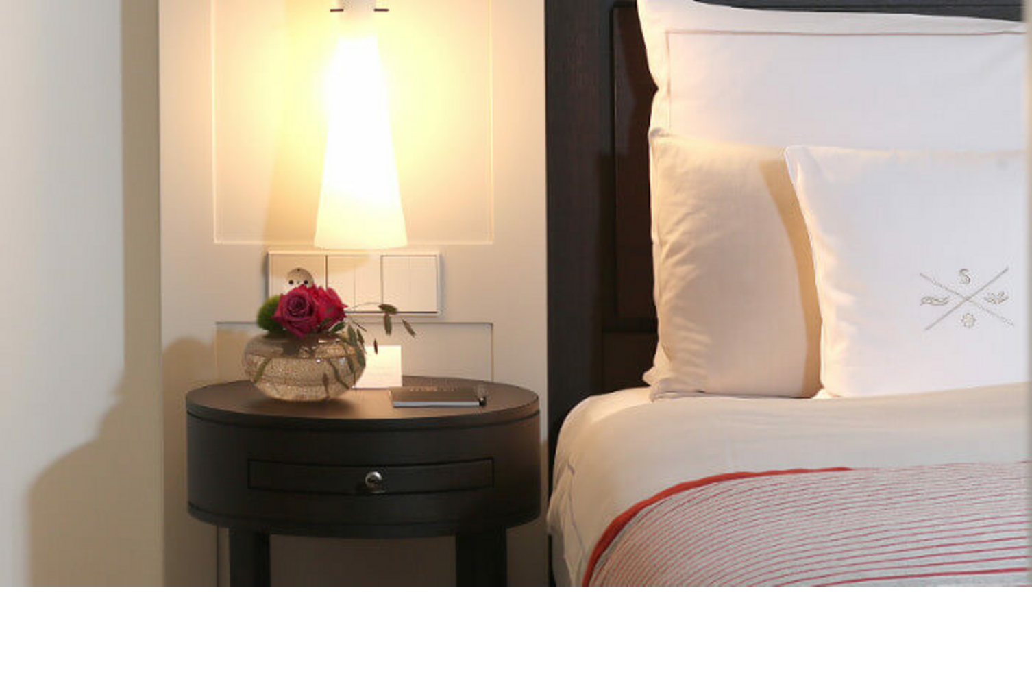 Bedside table with lamp, vase with rose and book next to elegant hotel bed with white pillows and striped comforter.