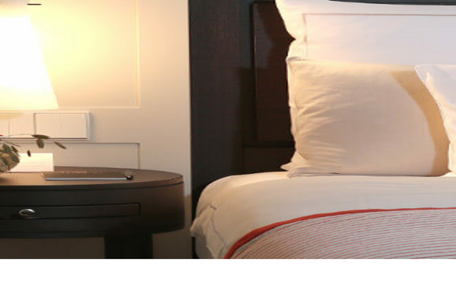 Nachttisch mit Lampe, Vase mit Rose und Buch neben elegantem Hotelbett mit weißen Kissen und gestreifter Bettdecke.