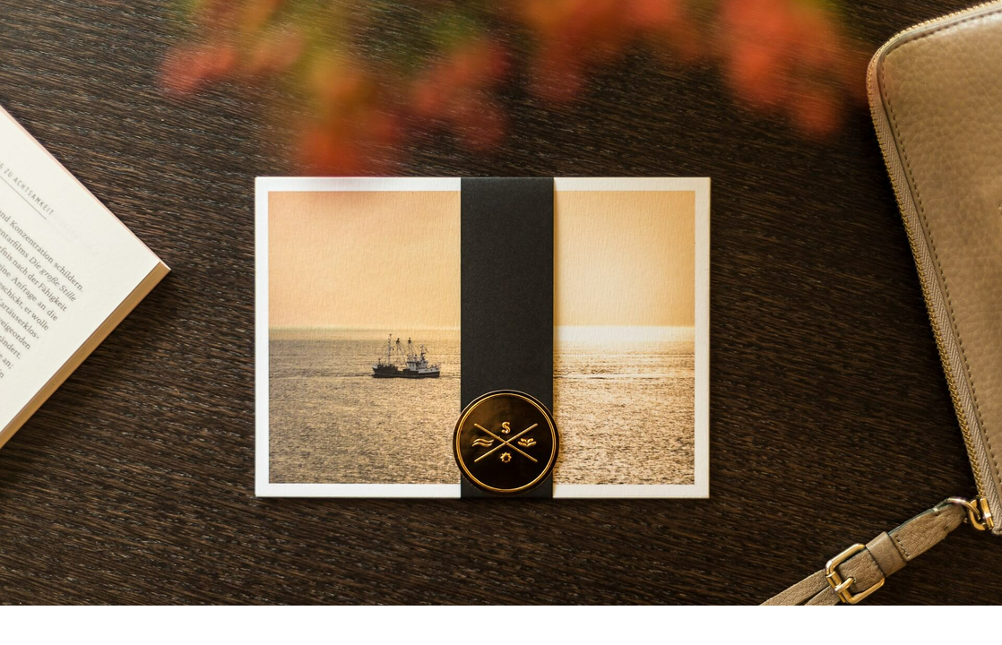 Postkarte mit Meerbild und Segelschiff, dekoriert mit schwarzem Band und goldenem Siegel, auf Holztisch.