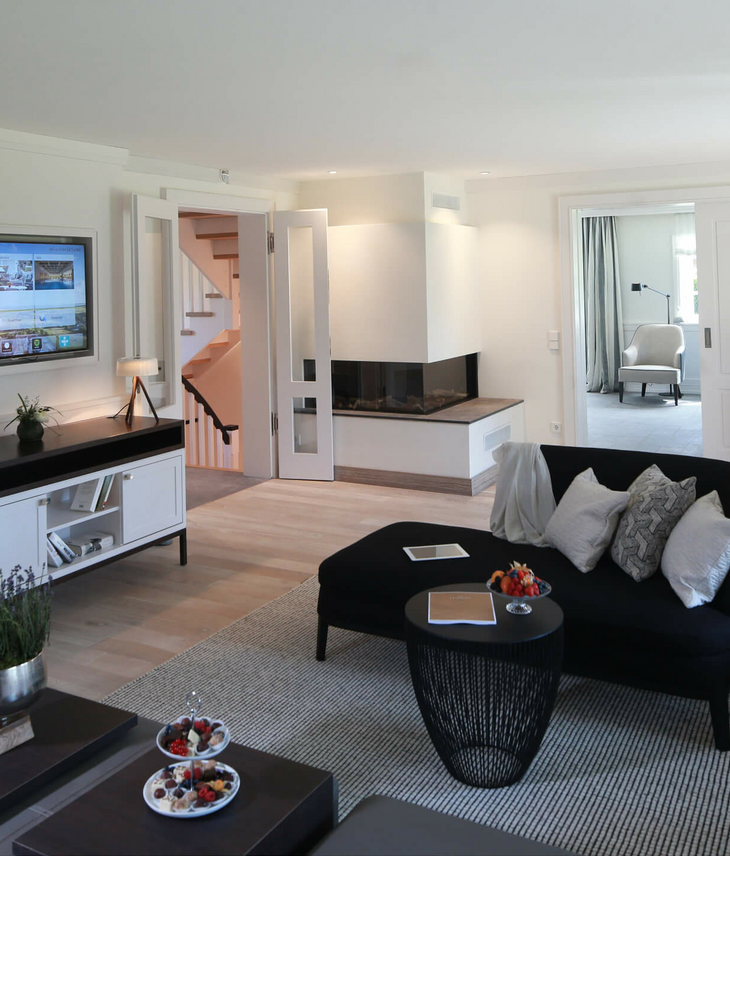 Wohnbereich der Villa Severin*s im Severin*s Resort und Spa auf Sylt Elegantes Wohnzimmer im Severin*s Resort & Spa mit Kamin, Sofa, modernem Dekor und Blick in ein helles Zimmer.