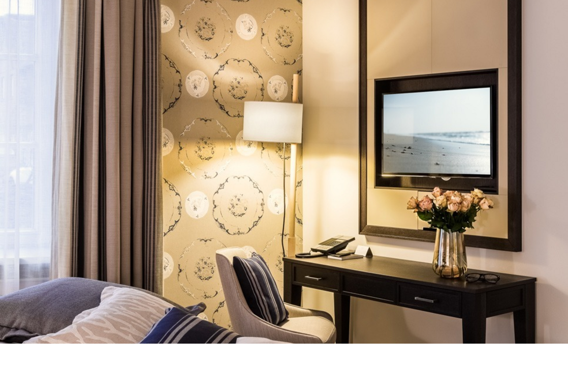 Elegantes Hotelzimmer mit gemütlichem Bett, stilvollen Kissen, Schreibtisch, Lampe und dekorativer Tapete.