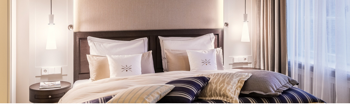 Elegantes Hotelzimmer mit großem Bett, dekorativen Kissen, stilvollen Nachttischlampen und bodenlangen Vorhängen.