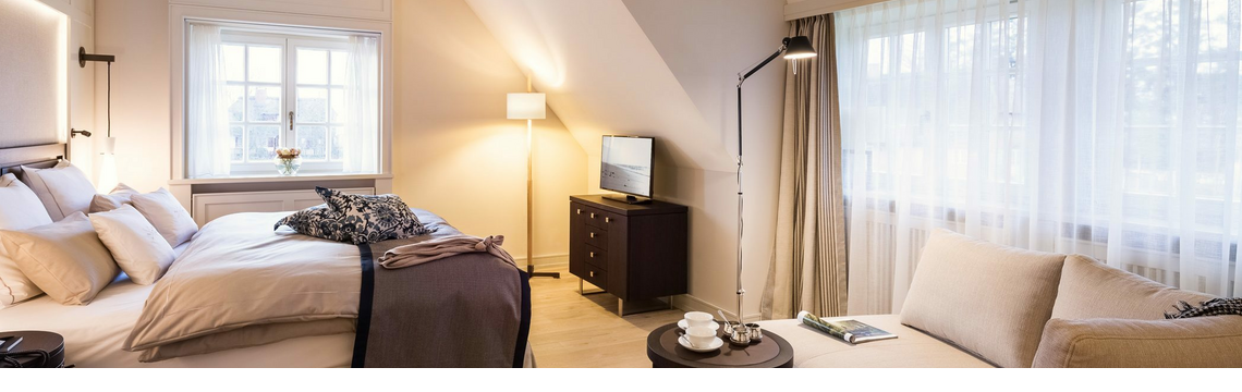 Helles, elegantes Hotelzimmer mit Doppelbett, Sofa, Tisch und Fernseher, große Fenster mit Vorhängen, gemütliche Atmosphäre.
