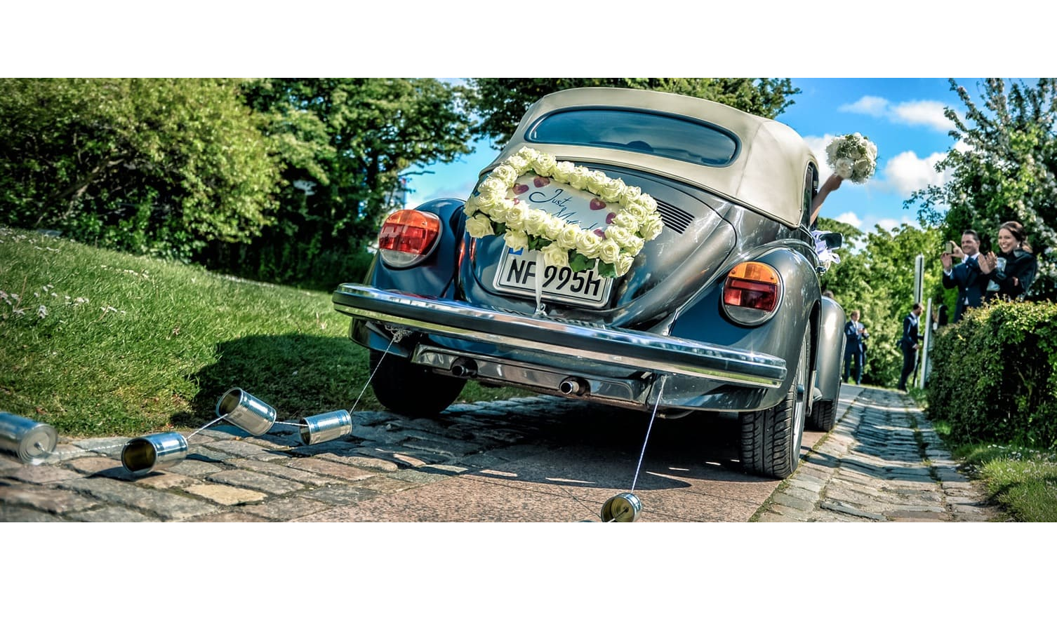 Wedding car with flower wreath and cans at the rear, driving on a paved road, surrounded by green landscape.