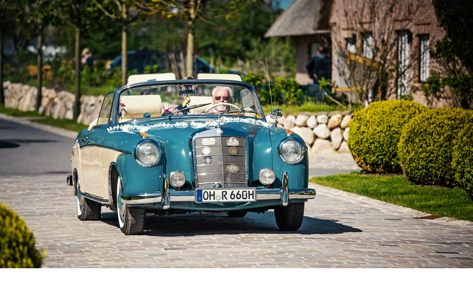 Classic convertible on cobblestones in front of a hotel, surrounded by green countryside.