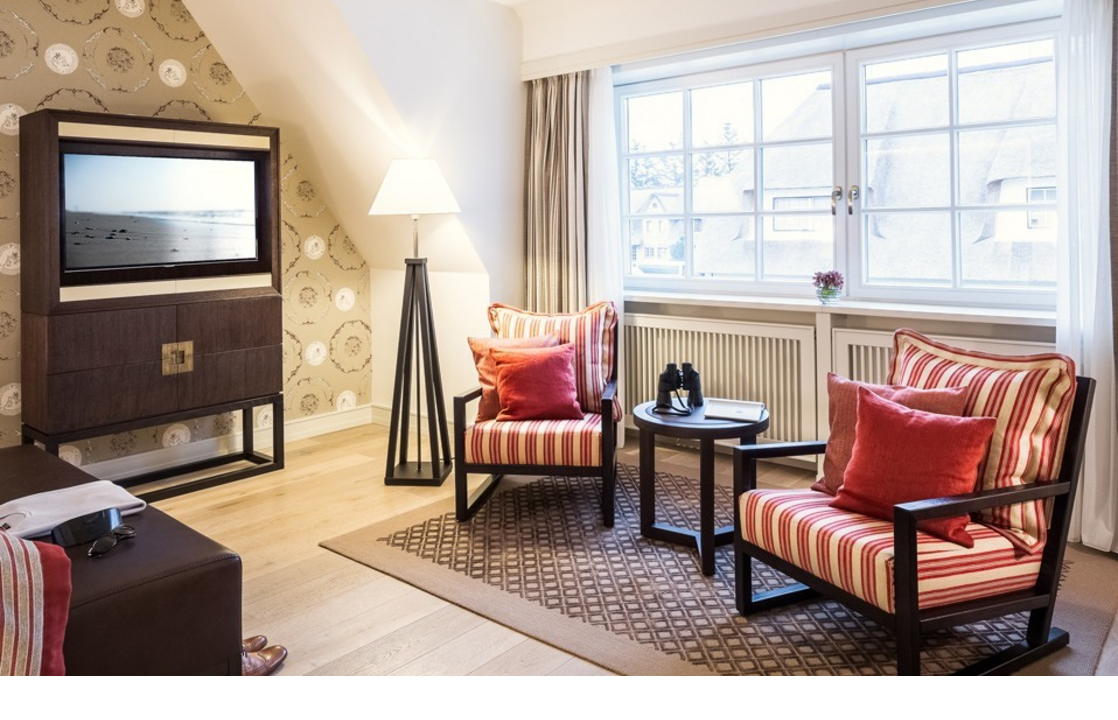 Bright living room with two striped armchairs, table, floor lamp and large window at Severin*s Resort & Spa, Sylt.