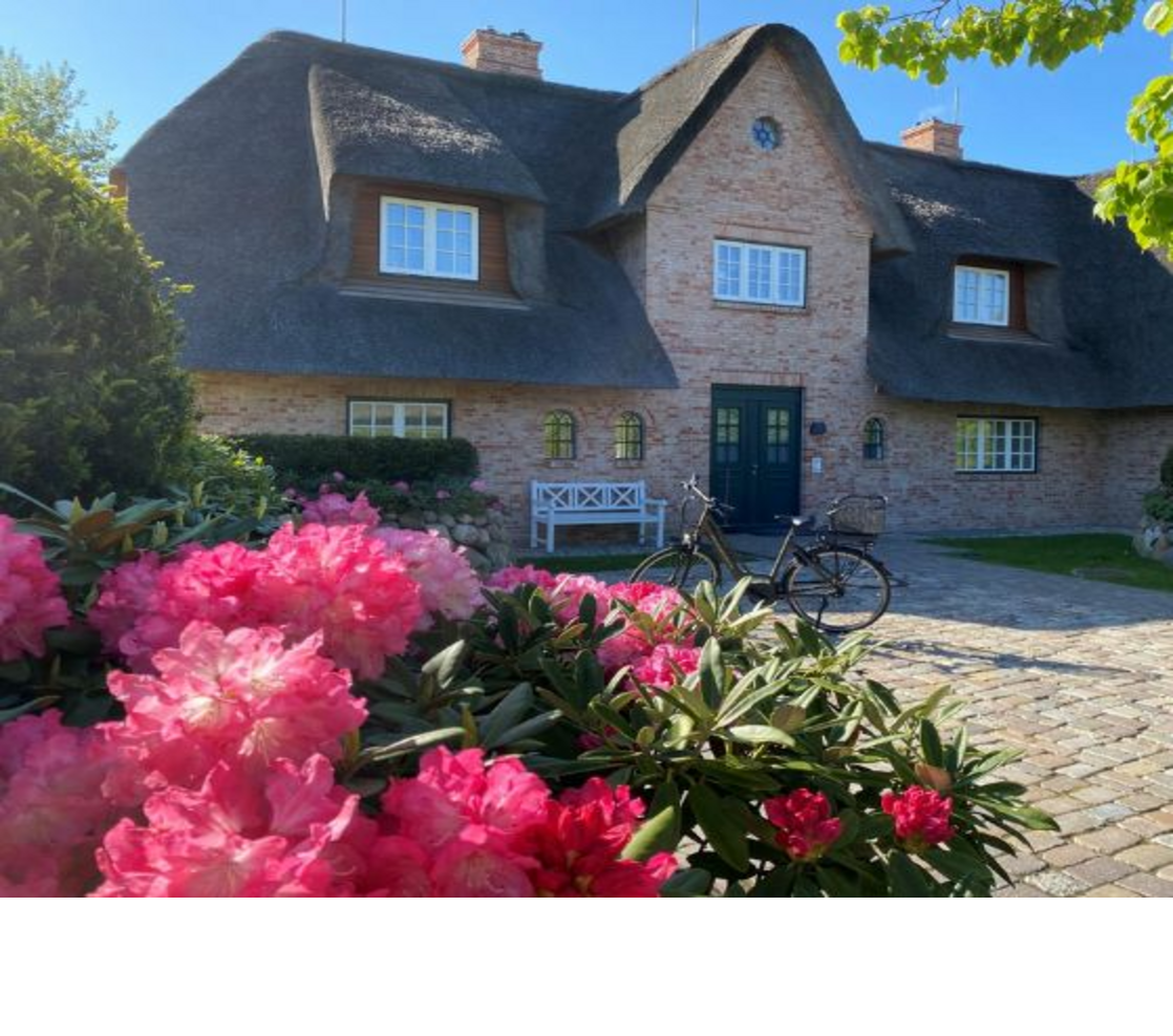 Reetgedecktes Haus mit blühenden Rhododendren und Fahrrad im Vordergrund, Teil des Severin*s Resort & Spa auf Sylt.